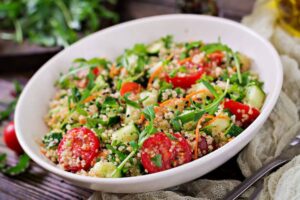 A vibrant quinoa salad featuring cherry tomatoes, cucumber, greens, and shredded carrots, served in a white bowl on a rustic table.