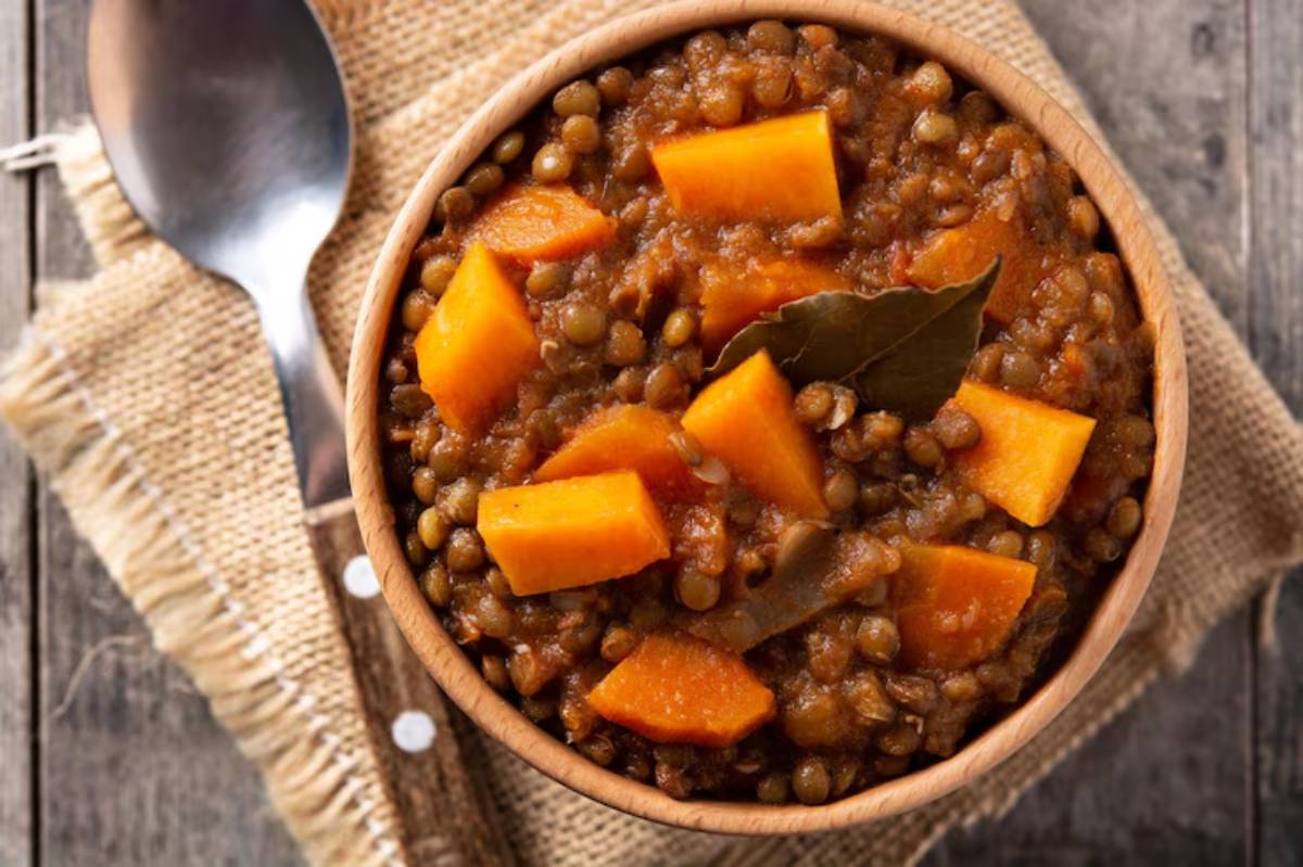 A wooden bowl filled with lentil stew, featuring chunks of orange sweet potato and bay leaves, on a burlap-covered surface.