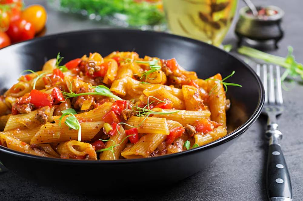 A black bowl filled with hearty pasta mixed with ground meat, tomatoes, and herbs, garnished with fresh greens.