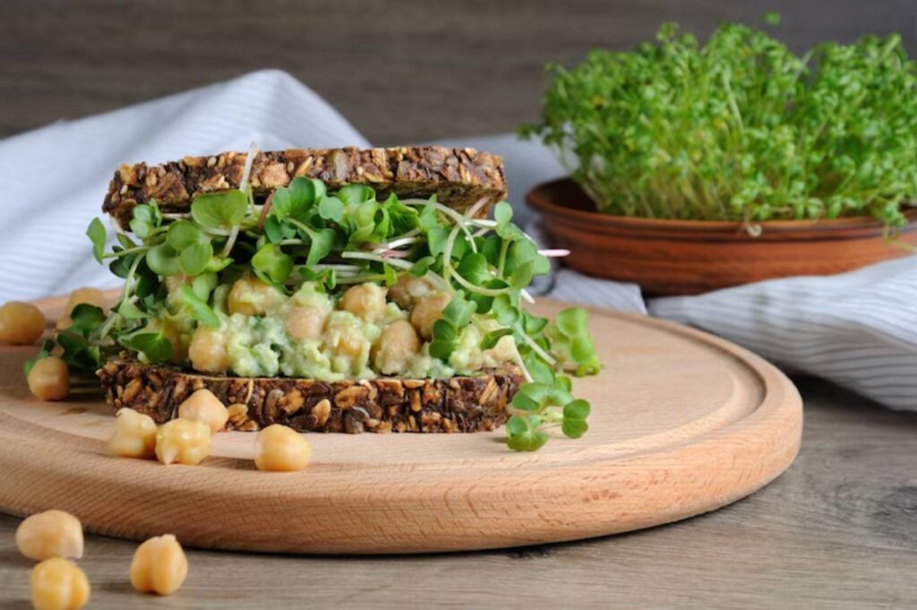 A nutritious sandwich layered with chickpeas, avocado, and sprouts on grainy bread, with chickpeas scattered on a wooden cutting board.