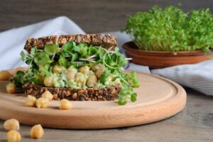 A nutritious sandwich layered with chickpeas, avocado, and sprouts on grainy bread, with chickpeas scattered on a wooden cutting board.