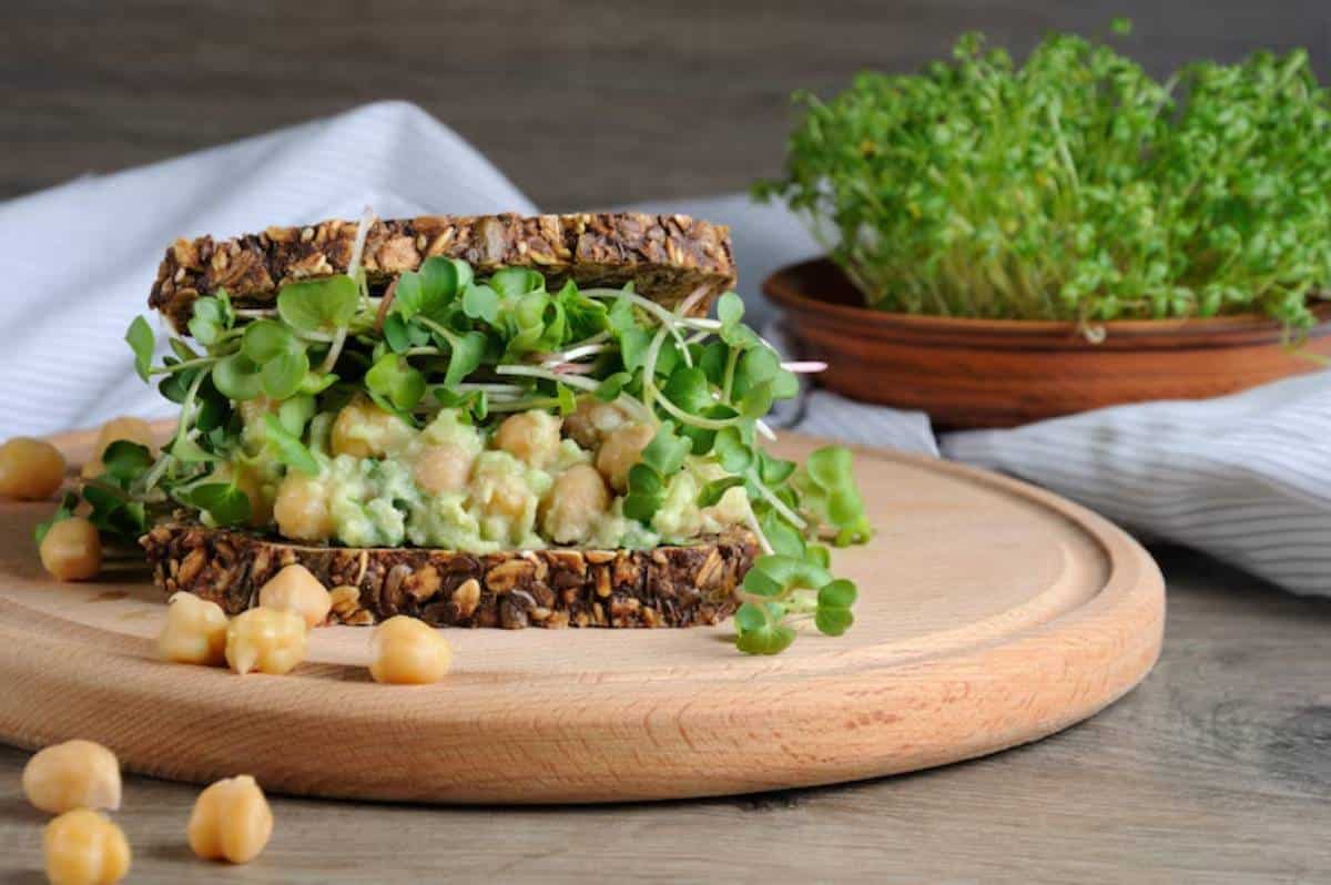 A nutritious sandwich layered with chickpeas, avocado, and sprouts on grainy bread, with chickpeas scattered on a wooden cutting board.