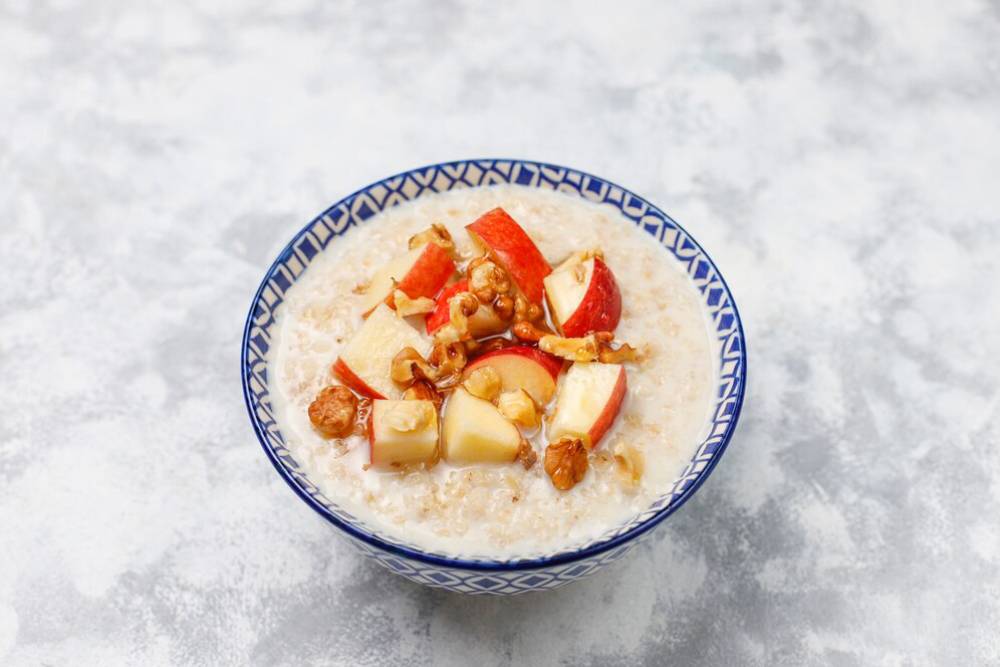 A bowl of oatmeal topped with sliced red apples and chopped walnuts on a textured gray background.