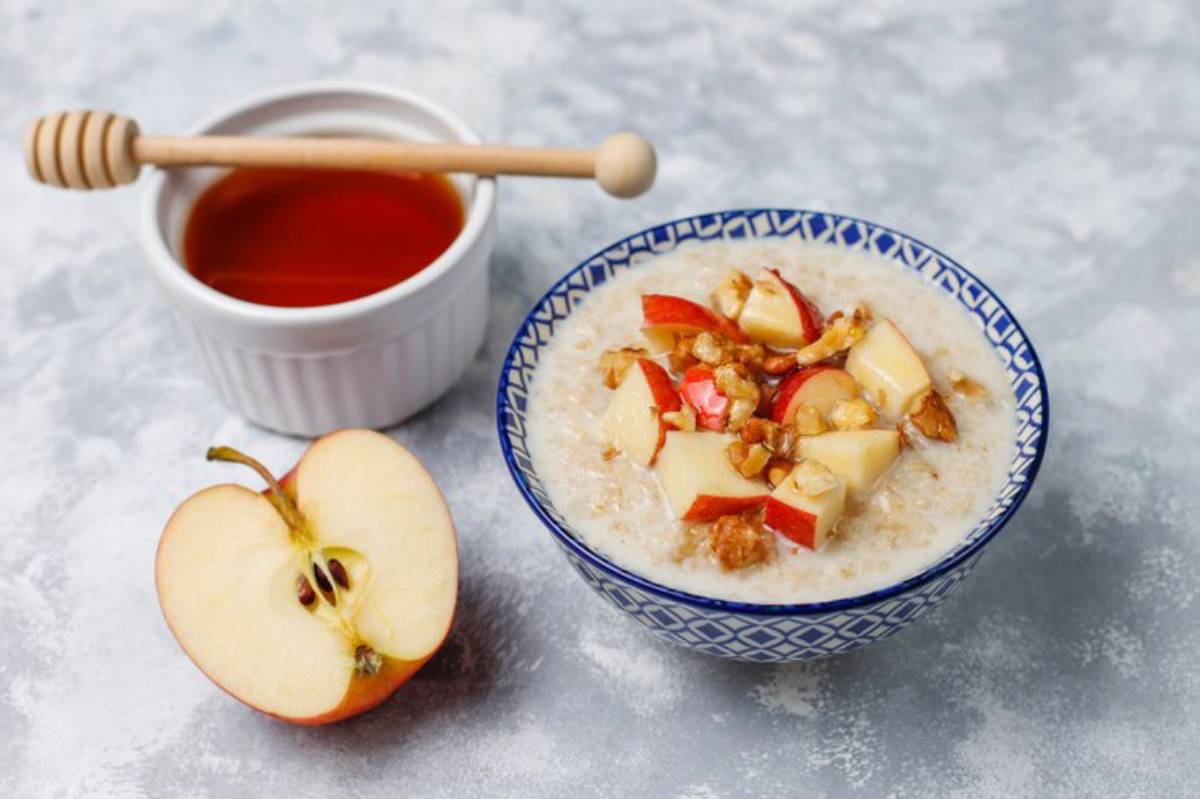 A bowl of oatmeal topped with chopped red apple and walnuts, served next to a white ramekin filled with honey and a wooden honey dipper, with a halved red apple placed nearby on a textured gray surface.
