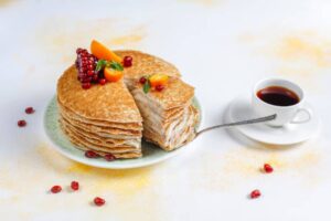 A layered pancake cake topped with berries and orange slices, served beside a cup of coffee on a white background.