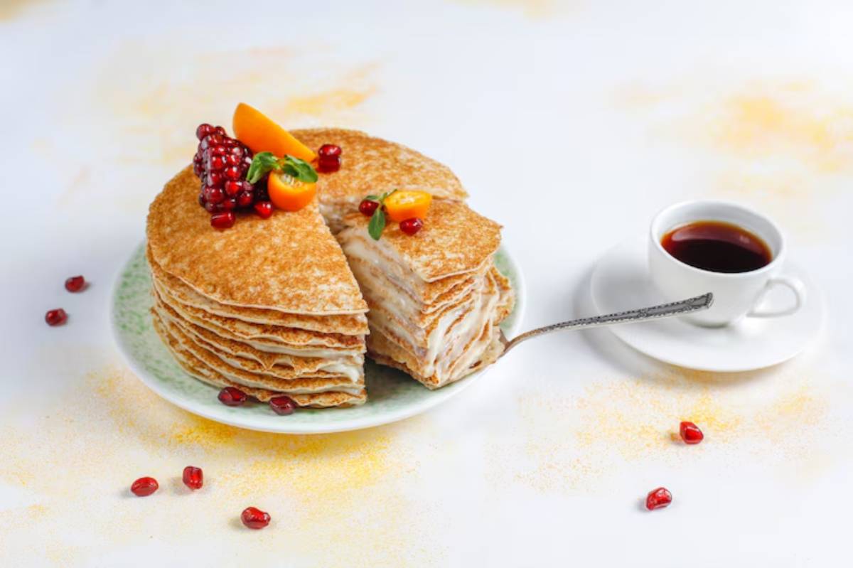 A layered pancake cake topped with berries and orange slices, served beside a cup of coffee on a white background.