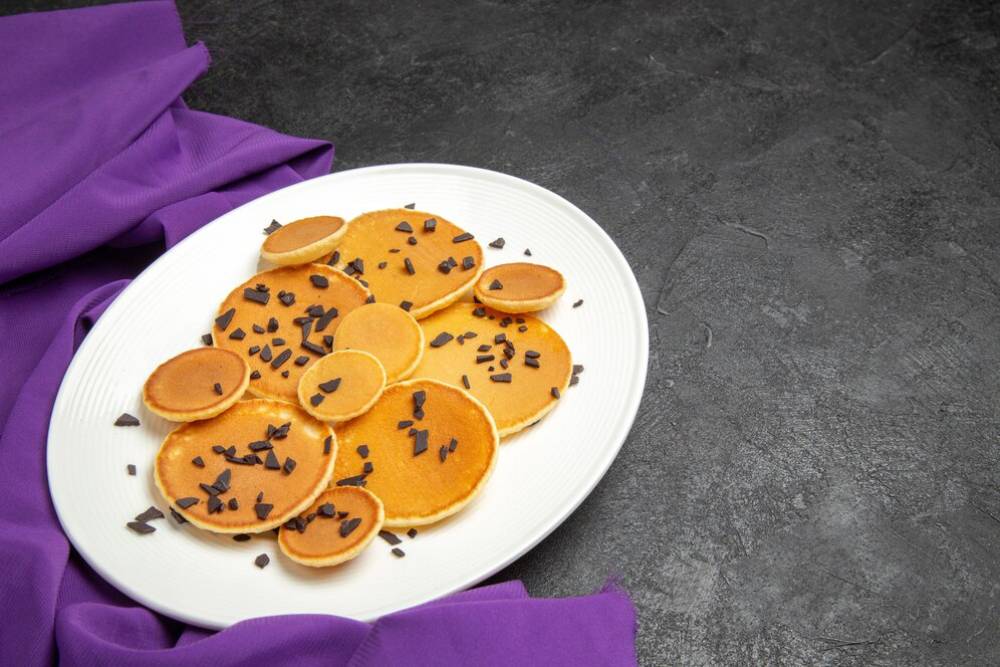 A plate of various-sized pancakes topped with chocolate shavings, placed on a dark surface with a purple cloth beside it.