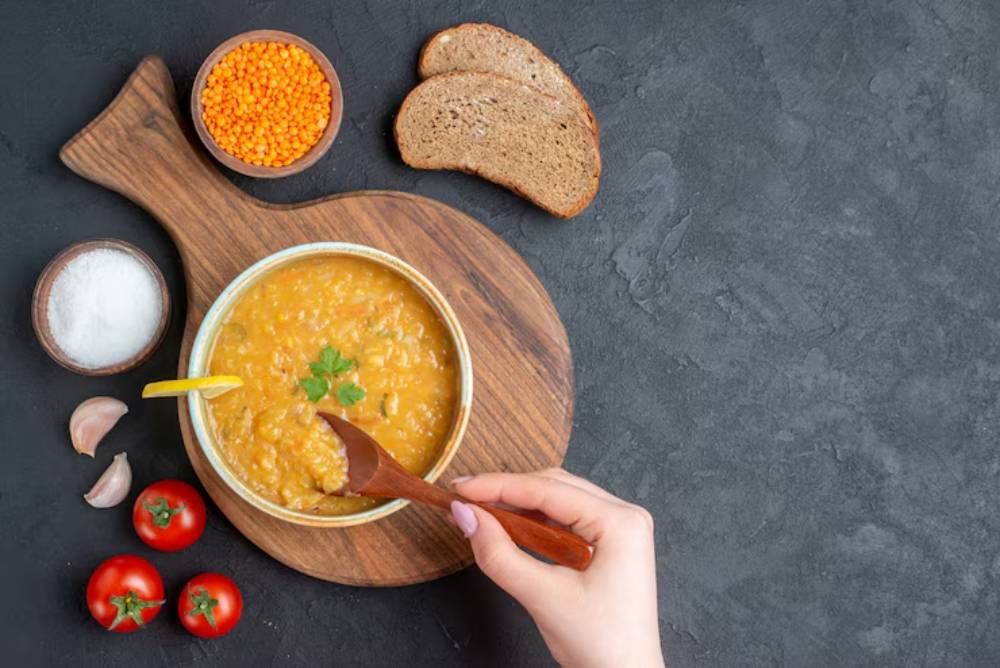 A hand with a wooden spoon stirs a bowl of yellow lentil soup on a wooden board, accompanied by bread, tomatoes, garlic, and spices.