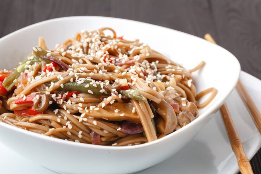 Close-up of a white bowl filled with noodles mixed with colorful vegetables and garnished with sesame seeds, chopsticks nearby.