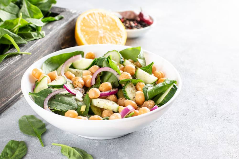 Fresh salad in a white bowl featuring spinach, chickpeas, cucumber, red onion, and lemon, with greens and spices in the background.