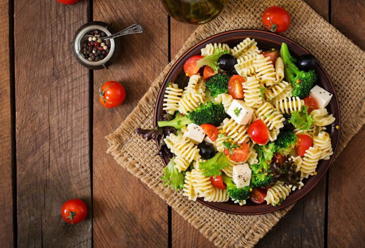 A vibrant pasta salad featuring rotini pasta, cherry tomatoes, broccoli, black olives, feta cheese, and fresh greens on a wooden table.
