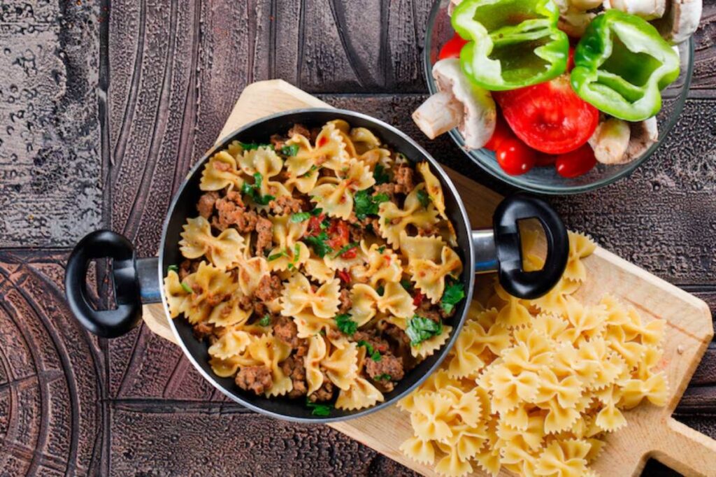 A pot of farfalle pasta mixed with minced meat and herbs, alongside a bowl of fresh vegetables including bell peppers and mushrooms.