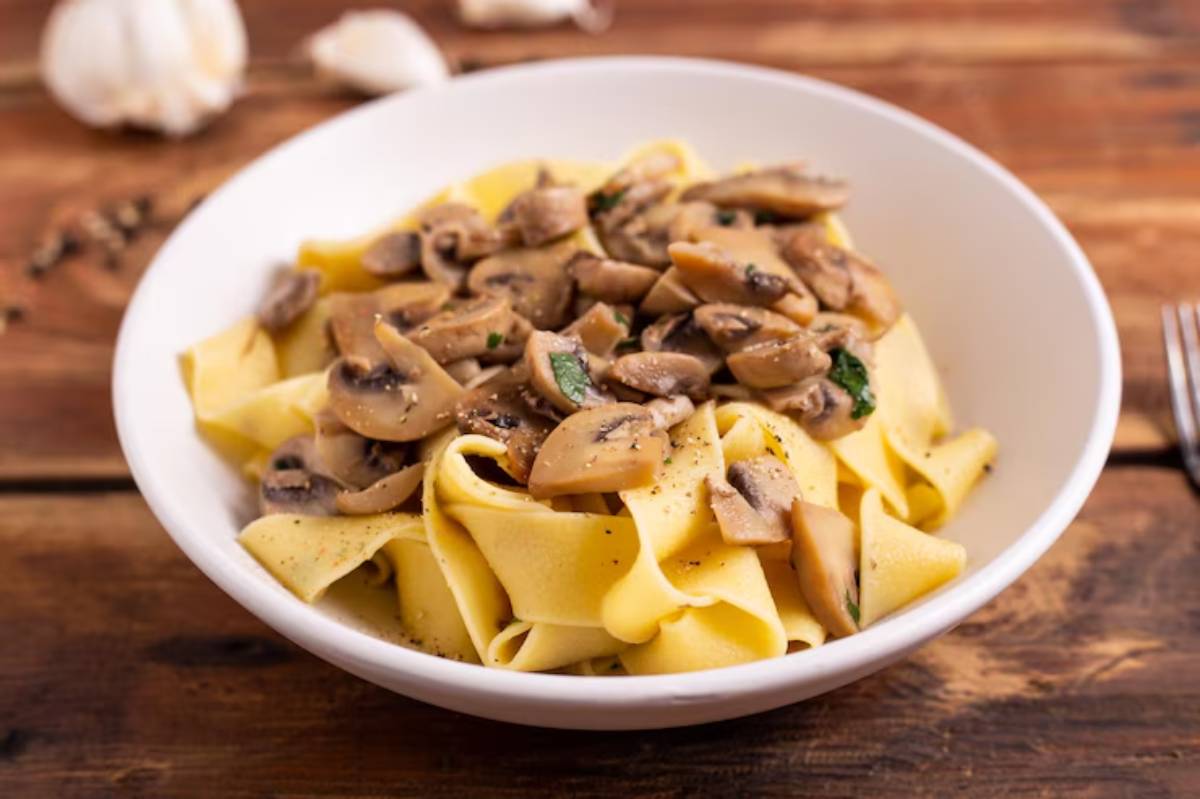 A bowl of pappardelle pasta topped with sautéed mushrooms and herbs, served on a rustic wooden table with garlic in the background.