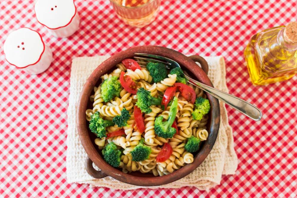 A bowl of pasta with broccoli and tomatoes on a checkered tablecloth, accompanied by salt shakers and olive oil.