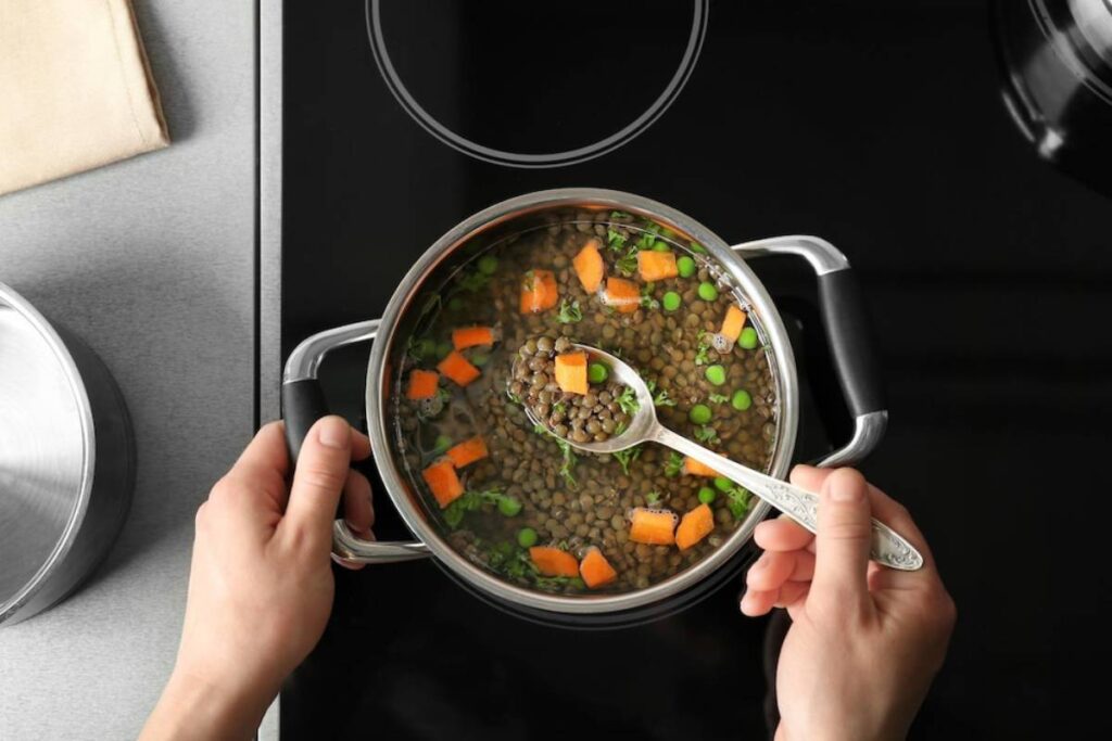A hand holding a spoon stirs a pot of lentil soup with carrots and peas on a sleek stovetop.