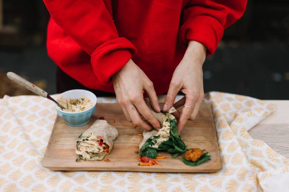A person in a red sweatshirt prepares a wrap filled with vegetables on a wooden board, next to a bowl of spread and a spoon.