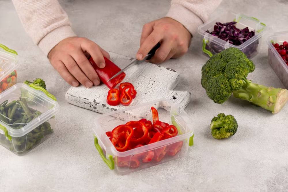 A person slices red bell peppers on a cutting board, surrounded by various containers of vegetables and a broccoli stalk on a gray surface.