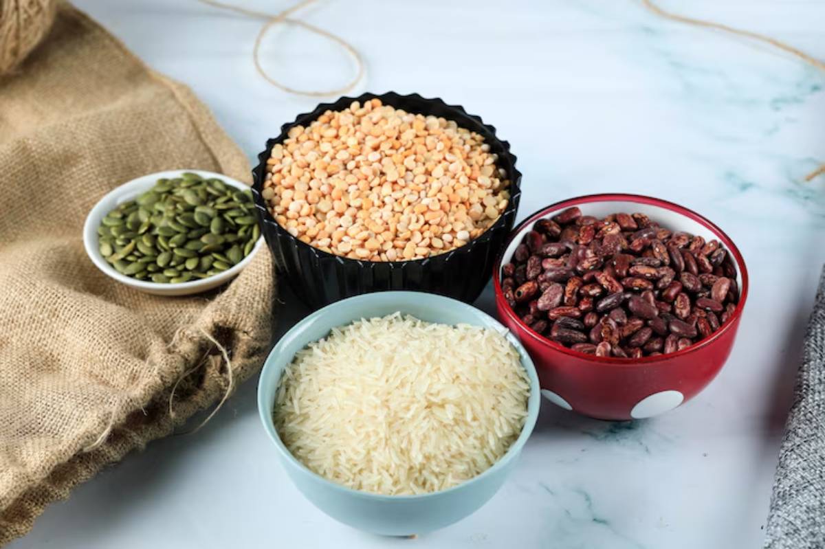Three bowls of food ingredients: yellow lentils, white rice, red beans, and green pumpkin seeds, with burlap fabric in the background.
