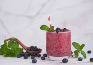 A vibrant blueberry smoothie in a glass garnished with mint leaves and a striped straw, surrounded by fresh blueberries and mint.