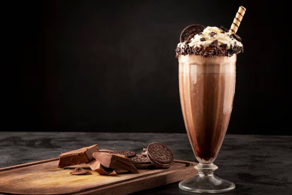 A chocolate banana smoothie in a glass, topped with chocolate shavings, sits beside fresh bananas and striped straws on burlap.