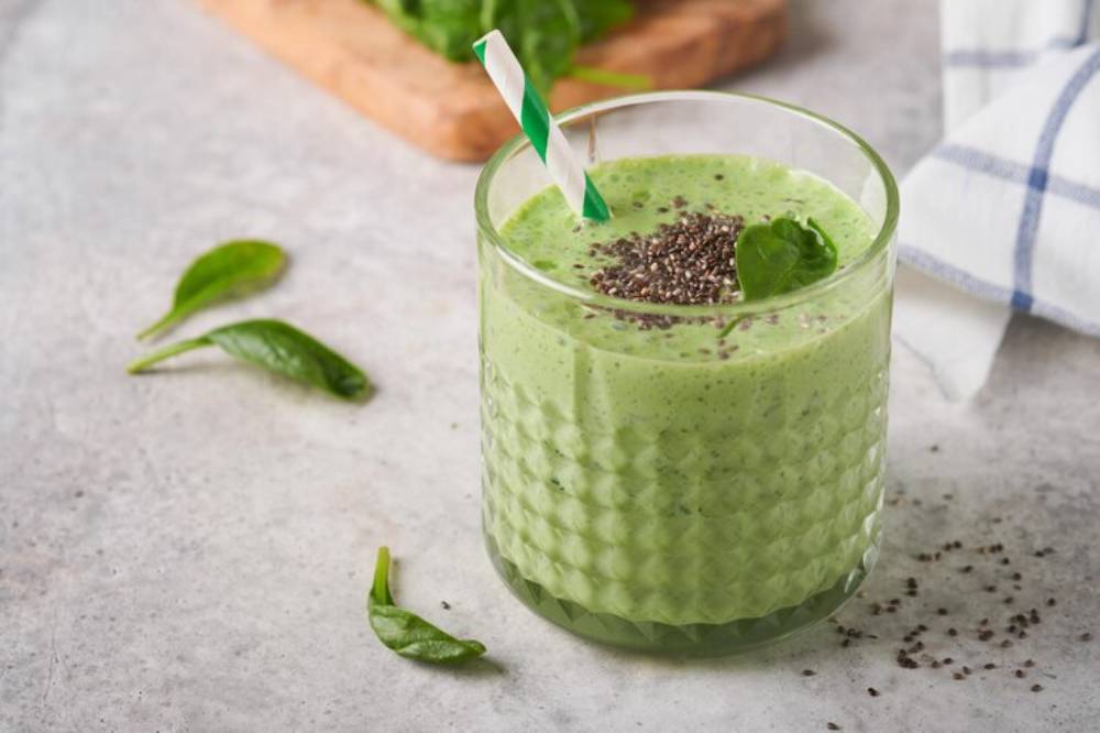 A refreshing green smoothie topped with chia seeds and a spinach leaf, served in a glass with a striped straw on a light countertop.