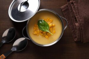 A pot of creamy corn soup garnished with a fresh spinach leaf, accompanied by two metal spoons, resting on a wooden table.