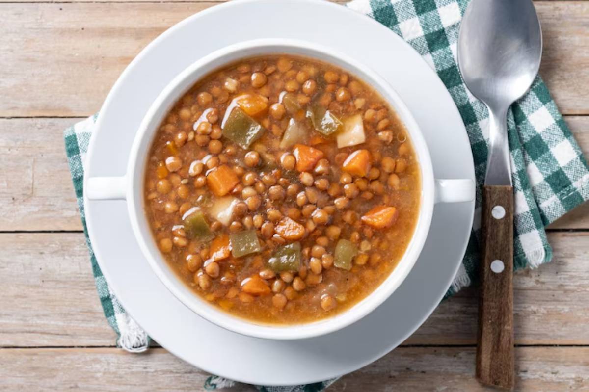 A bowl of hearty lentil soup with carrots, green bell pepper, and potatoes, served on a checkered napkin beside a spoon.
