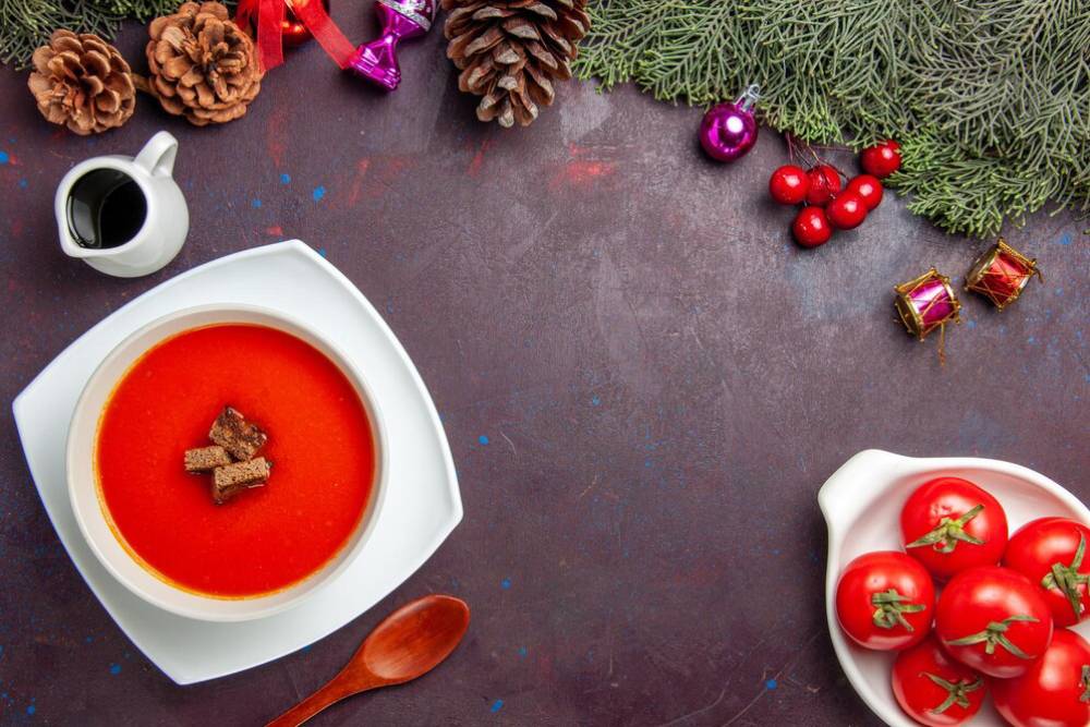A vibrant bowl of tomato soup topped with croutons, surrounded by festive decorations, pinecones, and fresh tomatoes.