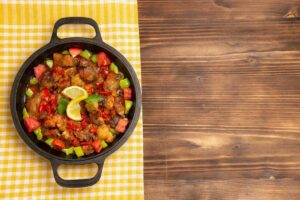 A cast iron skillet filled with colorful sautéed vegetables and tender meat, garnished with fresh lemon and herbs on a checkered tablecloth.