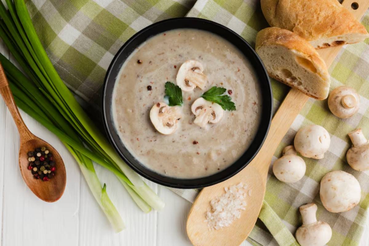 A bowl of creamy mushroom soup garnished with sliced mushrooms and herbs, accompanied by bread and fresh ingredients on a checkered cloth.
