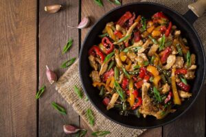 A colorful stir-fry in a black skillet, featuring chicken, bell peppers, and green beans, garnished with green onions and sesame seeds.