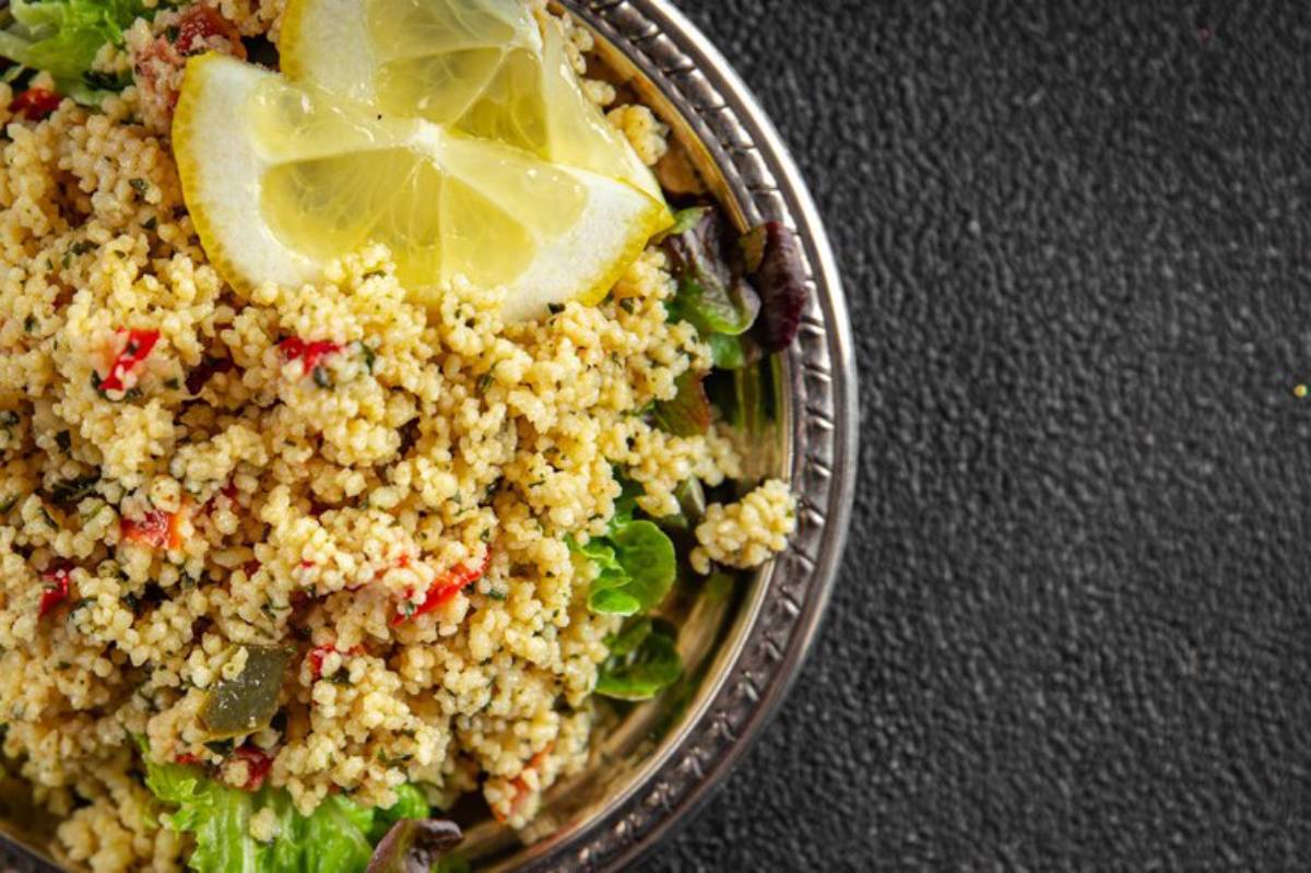 Couscous salad topped with lemon slices, mixed with vegetables and greens in a decorative silver bowl on a textured black surface.