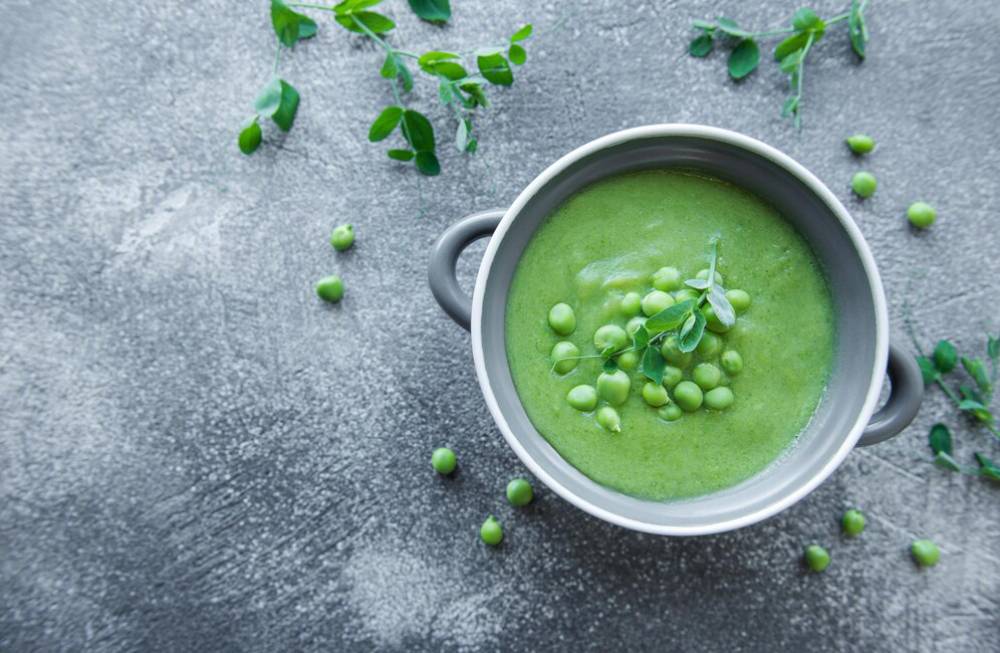 A bowl of vibrant green pea soup garnished with fresh peas and herbs, set against a textured gray background.