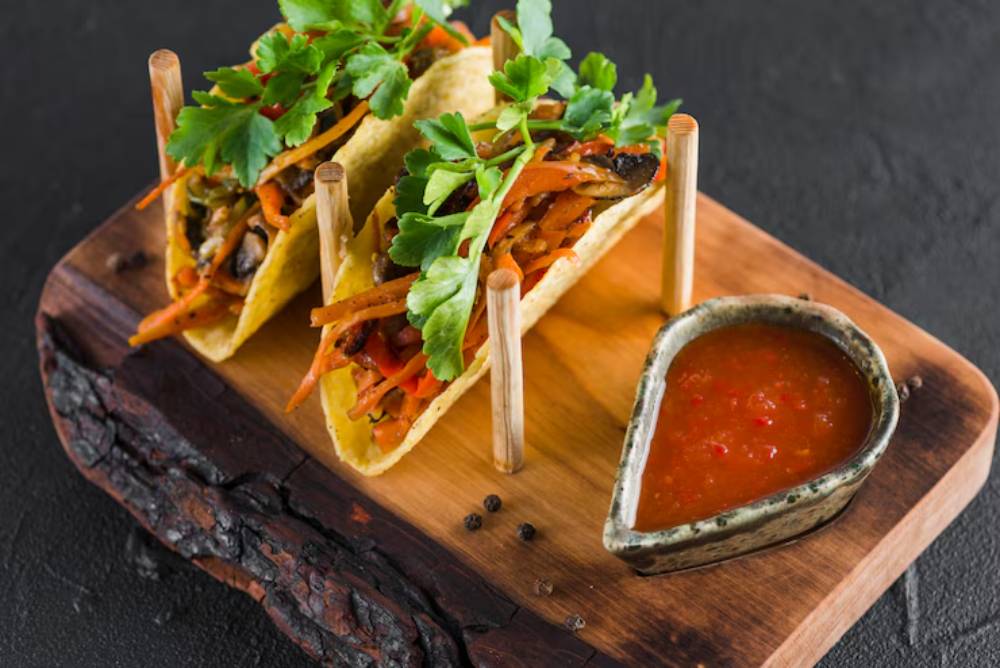 Two vibrant tacos filled with vegetables and topped with fresh herbs, served on a wooden board alongside a small dish of sauce.