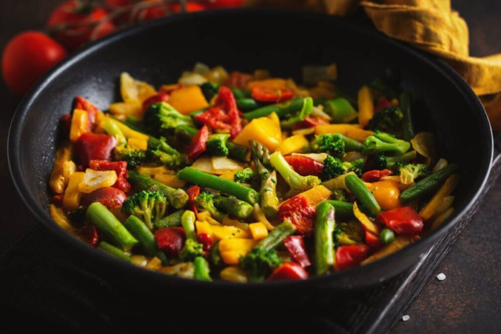 A colorful stir-fry of broccoli, bell peppers, green beans, and tomatoes in a black frying pan, garnished with fresh herbs.