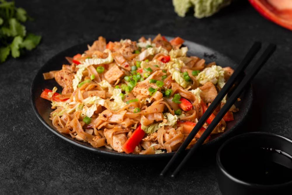 A delicious plate of stir-fried noodles with chicken, red peppers, and green onions, served with chopsticks and a side of soy sauce.