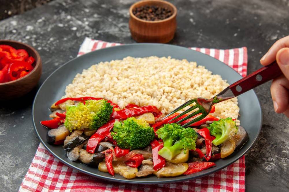 A hand holding a fork hovers over a plate of stir-fried vegetables and brown rice, with sliced red peppers in a bowl beside it.