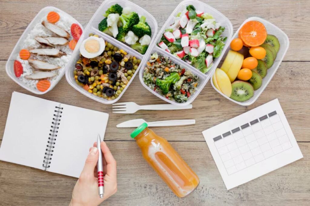 A hand holds a pen over a notebook beside healthy meal prep containers filled with grains, veggies, fruits, and a juice bottle.