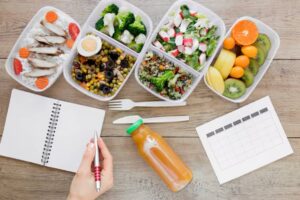 A hand holds a pen over a notebook beside healthy meal prep containers filled with grains, veggies, fruits, and a juice bottle.