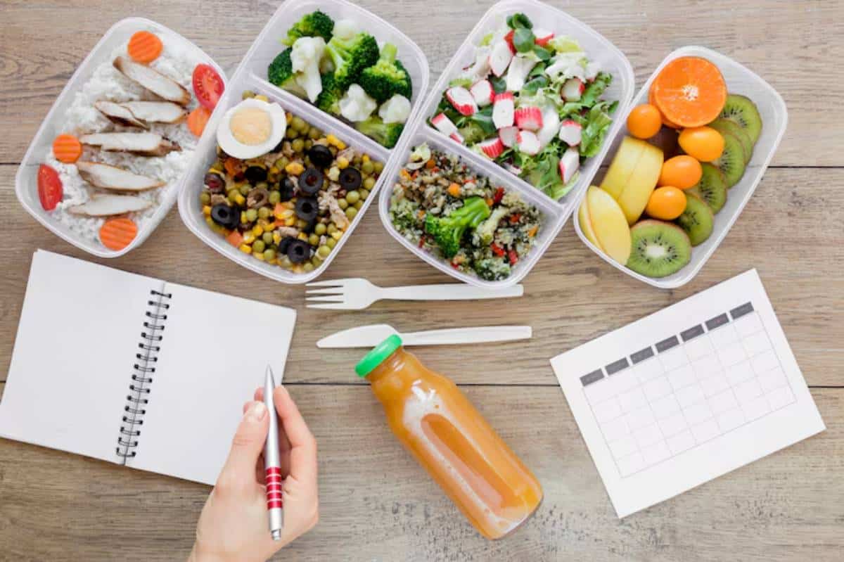 A hand holds a pen over a notebook beside healthy meal prep containers filled with grains, veggies, fruits, and a juice bottle.