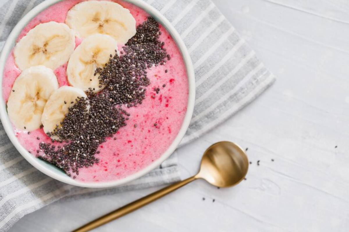 A vibrant smoothie bowl topped with banana slices and chia seeds, on a striped napkin beside a gold spoon.