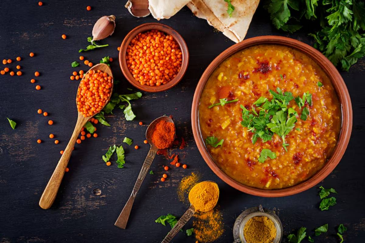 A wooden bowl filled with yellow lentil soup garnished with cilantro, surrounded by spices, orange lentils, and garlic cloves.