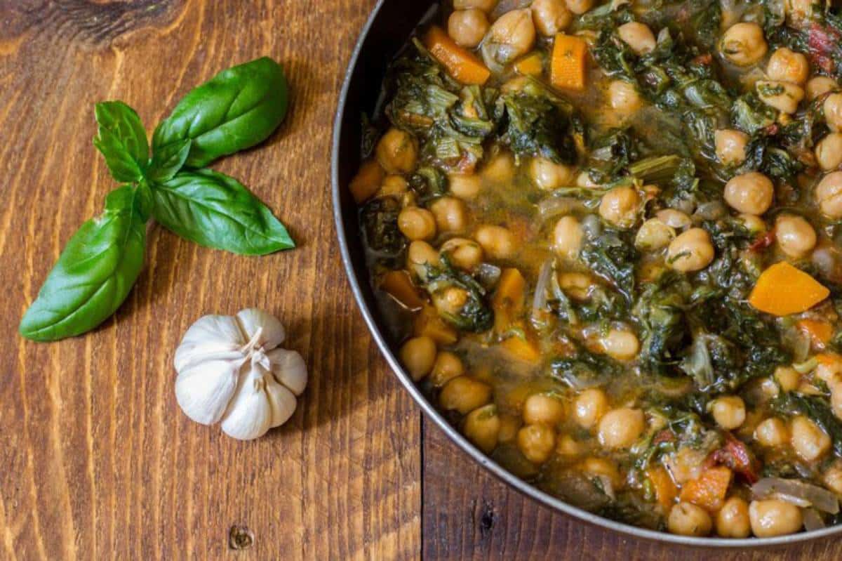 A bowl of cooked chickpea stew with greens and carrots, accompanied by fresh basil leaves and a garlic bulb on a wooden table.