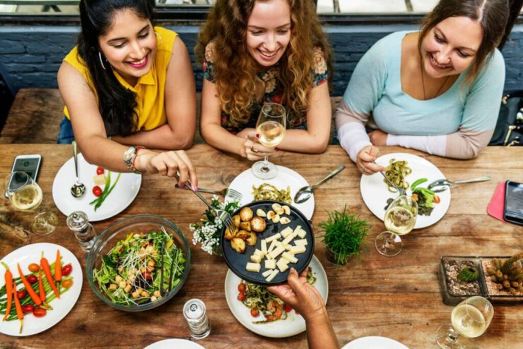 A diverse group of friends enjoys a meal at a wooden table, featuring salads, pasta, and a platter of cheese and roasted vegetables.