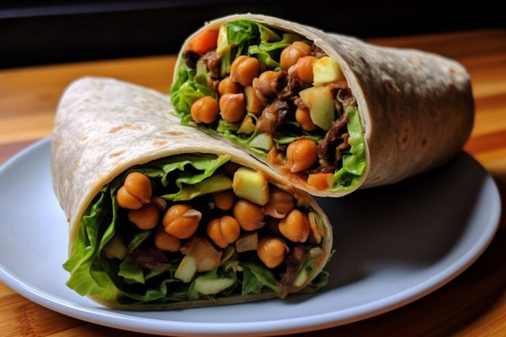 A close-up of two halved wraps filled with lettuce, chickpeas, diced apple, and meat, served on a white plate against a dark background.